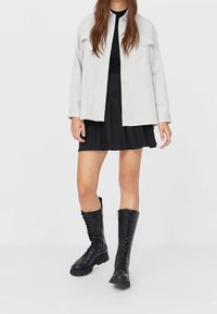 Lichtgrijze button-up shirtjack over een zwarte top en een geplisseerde zwarte rok, gecombineerd met zwarte lace-up laarzen tot de knie.