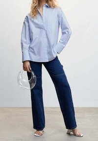 Lichtblauw gestreept overhemd met lange mouwen, gecombineerd met donkerblauwe wijde jeans en een doorzichtige ronde handtas. Witte hakken maken de look compleet.