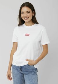 T-shirt en coton blanc avec un col rond et des manches courtes. Présente un texte brodé rouge qui dit "Doux et impossible à résister." Jeans en denim.