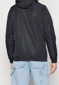 Czarna kurtka z kapturem wykonana z lekkiego materiału z elastycznym szwem u dołu. Posiada fakturowane wykończenie i kieszenie w szwach bocznych, noszona na jasnoniebieskich dżinsach.