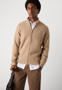 Beige ribbet zip-up cardigan i et mykt materiale, med høy krage og lange ermer, båret over en hvit stripete skjorte og brune bukser.