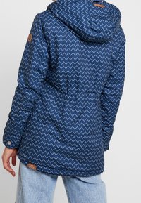 Polstret marineblå jakke med et zigzag-mønster i lyseblå. Har hætte, elastisk talje og brunt læderlogo-patch på siden.