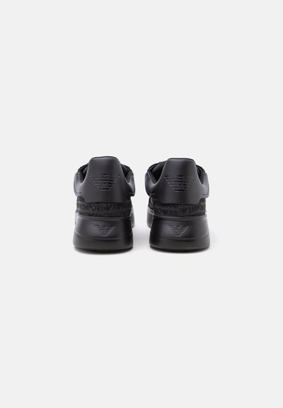 Paire de baskets noires avec logos en relief sur le talon et marquage autour de la bande supérieure du talon sur un fond clair uni.