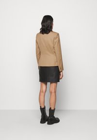 Blazer aderente beige con texture liscia, mini gonna nera di pelle e stivaletti neri, mostrati da dietro su uno sfondo neutro.