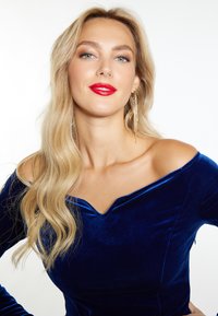Blå fløjl off-shoulder top med en dyb V-udskæring. Lange bølgete blond hår, rød læbestift og hængende guldøreringe. Glat tekstur.