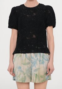 Top in pizzo nero con maniche a sbuffo presenta un motivo floreale in rilievo. Abbinato a pantaloni corti di colore chiaro con motivi di foglie di palma.