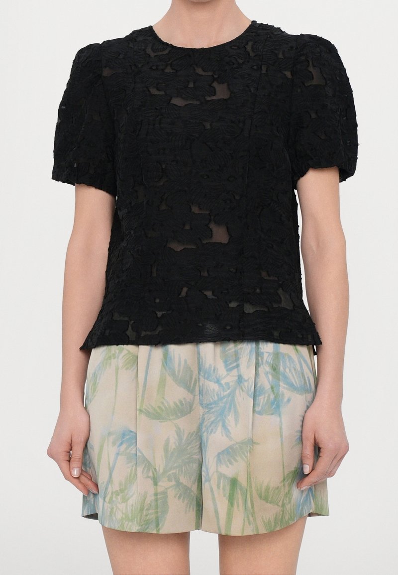 Top in pizzo nero con maniche a sbuffo presenta un motivo floreale in rilievo. Abbinato a pantaloni corti di colore chiaro con motivi di foglie di palma.