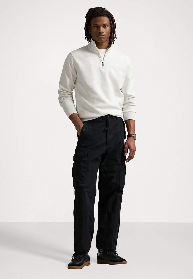 Felpa grigia chiara con zip e polsini a costine abbinata a pantaloni cargo neri con tasche laterali e polsini affusolati. Indossata con sneaker nere.