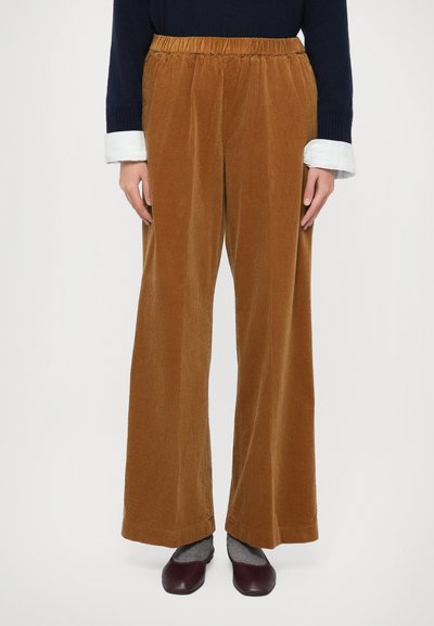 Personne portant un pantalon en velours côtelé large marron, des chaussettes grises, des chaussures à faible talon bordeaux, et un pull bleu marine avec des poignets de chemise blancs visibles.