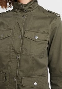 Olivově zelená bunda vyrobená z odolné tkaniny, s vysokým límcem, patentkami a čtyřmi předními kapsami s kovovými akcenty.