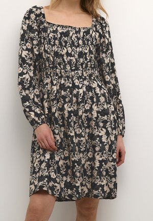 Frau trägt ein schwarzes, knielanges Kleid mit Blumenmuster, langen Puffärmeln und einem gerafften quadratischen Ausschnitt, steht vor einem schlichten Hintergrund.