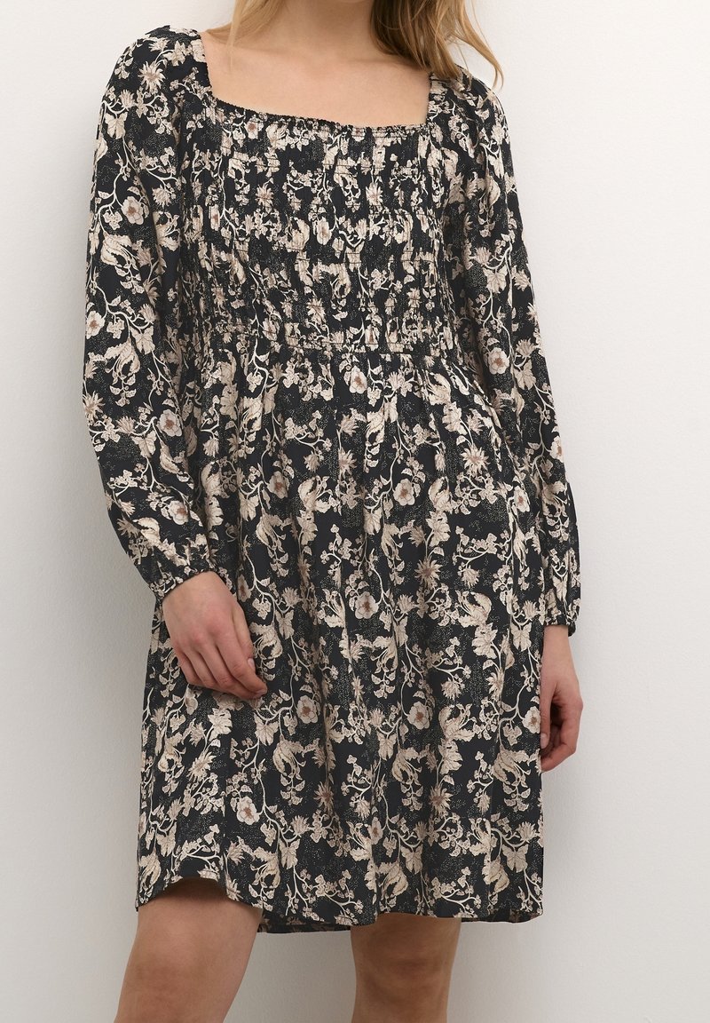 Femme portant une robe noire fleurie longueur genou avec des manches longues bouffantes et un col carré smocké, debout devant un fond uni.
