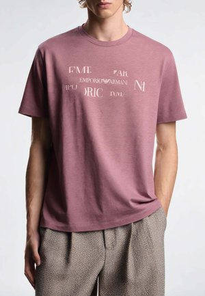 Giovane uomo con una T-shirt malva sbiadita con testo misto del logo Emporio Armani, abbinata a pantaloni beige texturizzati, in piedi davanti a uno sfondo semplice.