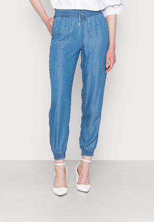 Woman wearing light blue elastic-waist jogger pants paired with white ankle-strap high heels and a white blouse with scalloped sleeves.