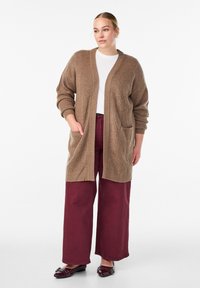 Cardigan marrone a maglia con una vestibilità rilassata, maniche lunghe, apertura frontale e due tasche anteriori, abbinato a pantaloni ampi color bordeaux e scarpe.