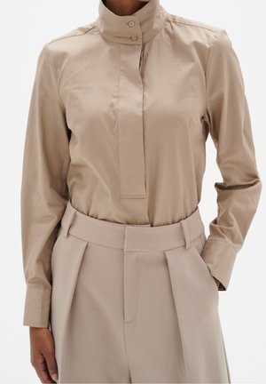 Persoon draagt een beige hooggesloten blouse met knopen, ingestopt in een bijpassende hoog getailleerde broek met riem en zijzakken.