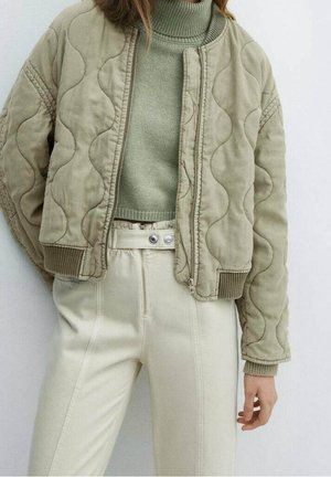 Mujer con chaqueta bomber acolchada verde sobre un suéter de cuello alto verde y pantalones crema de talle alto con cierre de doble botón.