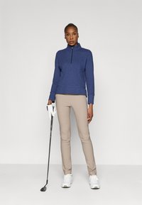 Sudadera de cremallera azul marino con cuello simulado, combinada con pantalones beige ajustados. El modelo sostiene un palo de golf, vistiendo zapatillas blancas y un guante blanco.