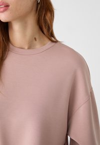 Helles rosa Sweatshirt mit weicher Textur, Rundhalsausschnitt und lockerer Passform. Verfügt über fallende Schulterabnäher und minimale Nähdetails.