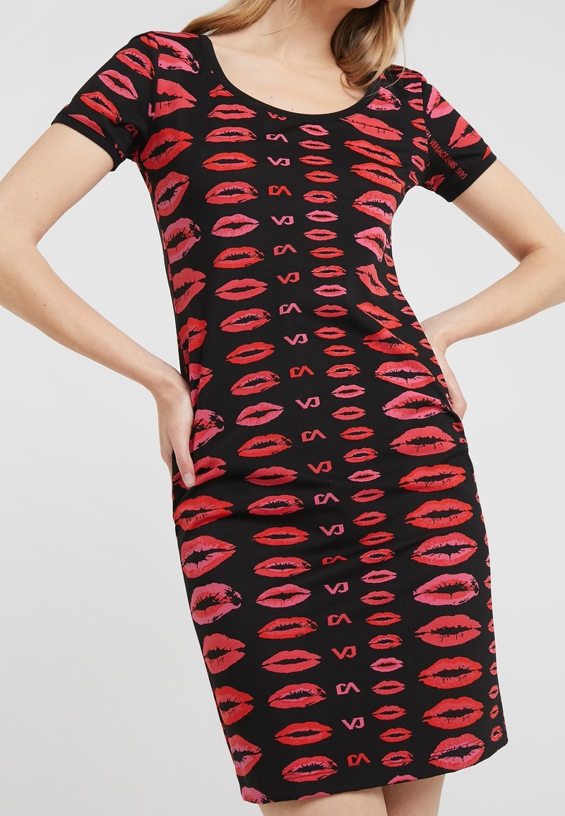 Femme portant une robe noire ajustée avec des motifs d'empreintes de lèvres rouges et roses et des manches courtes, posant avec les mains sur les hanches.