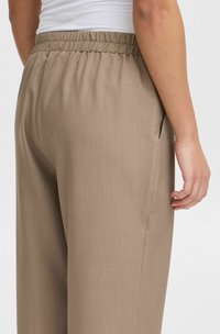 Pantalons lâches de couleur marron clair avec une taille élastique. Comprennent des poches latérales et un tissu lisse et texturé. Vue arrière présentée.