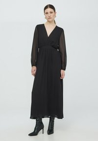 Abito maxi nero con maniche lunghe trasparenti, scollatura a V, vita aderente e gonna fluente. Abbinato a stivali neri con tacco alti fino al ginocchio.