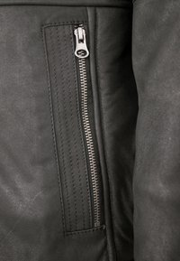 Schwarze Lederjacke mit einer Reißverschlusstasche. Die Textur ist glatt und weist gestickte Details sowie einen metallischen Reißverschluss auf.