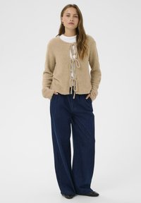 Smėlio spalvos tekstūruotas kardiganas su raišteliu priekyje, dėvimas ant baltos marškinės ir derinamas su tamsiai mėlynomis platėjantėmis kelnėmis bei juodais batais.