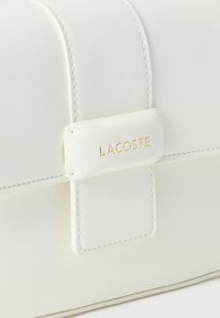 Λεπτομέρεια κλιπ τσάντας από λευκό δέρμα με ραμμένες άκρες και χρυσή ανάγλυφη επιγραφή "LACOSTE" στο λουράκι.