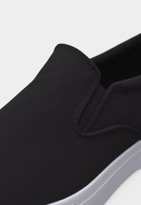 Chaussure noire en toile à enfiler avec panneau latéral élastique et semelle en caoutchouc blanche, présentée en gros plan.