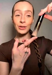 Frau mit geflochtenem Haar hält und zeigt auf eine Tube Abdeckstift, trägt ein braunes Oberteil vor schlichten Hintergrund.
