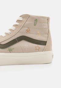Baskets montantes beiges avec lacets blancs, bande latérale vert olive et petits motifs brodés comprenant un cœur, une étoile, un soleil et des plantes.