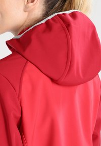 Rød vandtæt jakke med en tætsiddende hætte, glat tekstur og kontrasterende grå detaljer langs hættens kant.