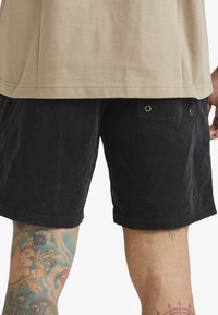 Shorts en velours côtelé noirs avec une poche arrière dotée d'œillets en métal ; portés avec un t-shirt beige. Tatouage complexe visible sur la jambe.