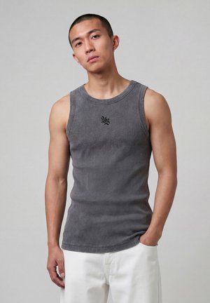 Joven con el cabello corto, vestido con una camiseta gris sin mangas con un pequeño logo negro y pantalones blancos, de pie frente a un fondo sencillo.