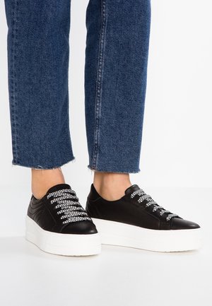 Czarne sneakersy na platformie z białymi sznurówkami w wzory, noszone z krótkimi niebieskimi dżinsami na białym tle.