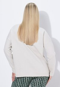 Eine blonde Frau, die mit dem Rücken zur Kamera steht, trägt einen lockeren beigen Pullover und schwarze Hose mit weißem Muster, vor einem schlichten weißen Hintergrund.