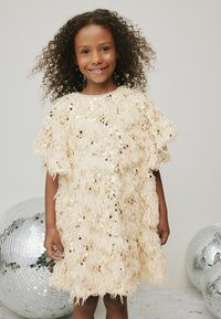 Rochie crem, cu maneci scurte, din pene, decorată cu paiete circulare. Croi lejer, până la genunchi, cu o textură distinctă. Copil îmbrăcat stă lângă bile disco.