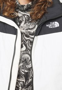 Biała puchowa kurtka z czarnymi akcentami, z wysokim kołnierzem i zamkiem z przodu, noszona na wzorze czarno-białego marmurkowego golfu.