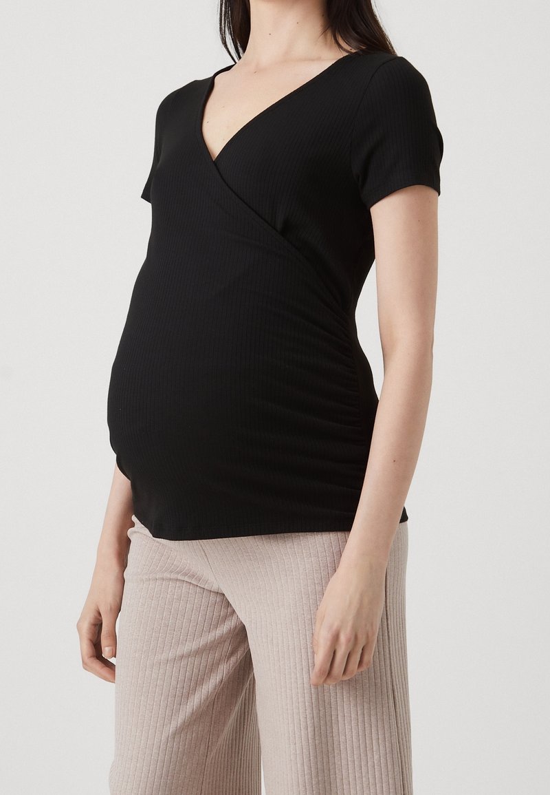 Femme enceinte portant un haut cache-cœur noir à manches courtes et un pantalon côtelé beige clair, debout devant un fond uni.