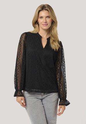 Black blouse with long lace sleeves featuring diamond patterns,  V-neckline, and ruffled cuffs, paired with light gray jeans.
