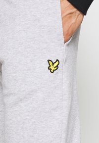 Ręka w kieszeni jasnoszarych spodni, widać mały żółty haftowany emblemat ptaka blisko szwu kieszeni.