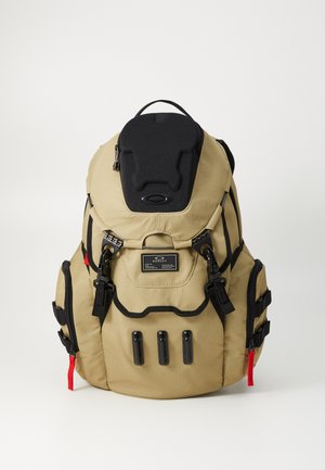 Tan backpack with black accents, durable fabric, padded top, multiple side pockets, and three black hardware loops on the front. Red zipper pull.