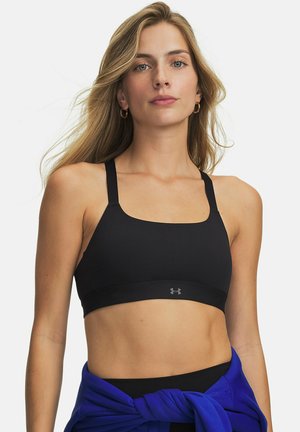 Junge Frau mit langen blonden Haaren, die einen schwarzen Sport-BH und eine blaue Jacke, um die Taille gebunden, vor weißem Hintergrund trägt.