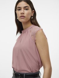 Blusa senza maniche rosa con dettagli in pizzo sulle spalle, caratterizzata da un colletto alto e morbide pieghe. Indossata con una cintura nera. Tessuto dalla texture liscia.