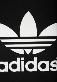 Mustat puuvillavaatteet, joissa on suuri valkoinen Adidasin trefoil-logo, jossa on kolme lehtimuotoa ja kolme vaakaraitaa pohjassa.