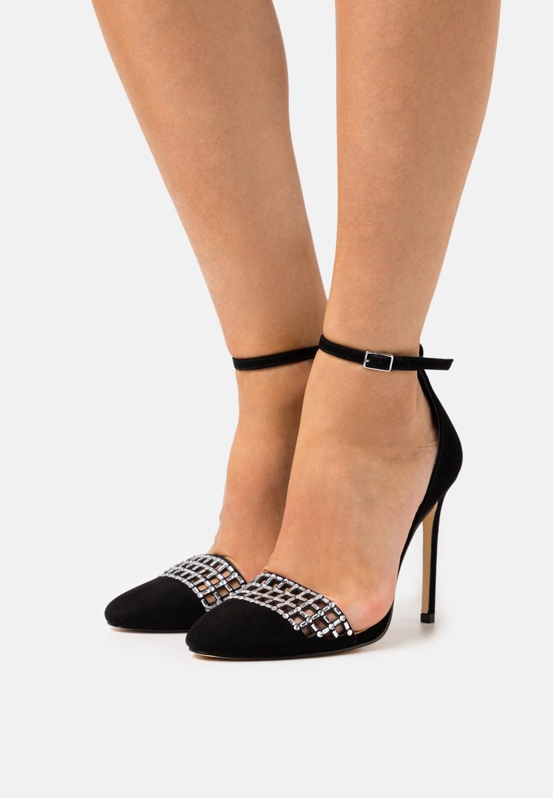 Jambes portant des chaussures à talons hauts noires avec brides à la cheville et détail en grille de cristaux argentés sur les bouts pointus, sur fond blanc.