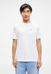 Λευκό βαμβακερό μπλουζάκι κοντό μανίκι, με στρογγυλή λαιμόκοψη και μικρή μαύρη λεπτομέρεια λογότυπου στην αριστερή πλευρά του στήθους, φορεμένο με ανοικτού γαλάζιους τζιν.