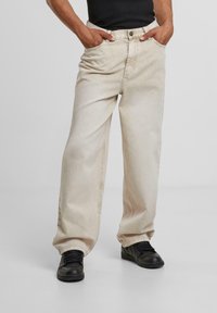 Beige wide-leg jeans made of denim, featuring a classic button and zipper fly, and side pockets. Worn with black sneakers.