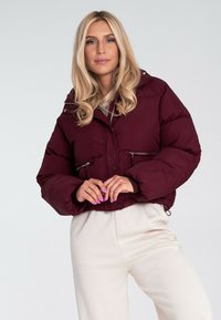 Bordowa kurtka puffer z krótki krojem, podwójnymi zamkami błyskawicznymi na przednich kieszeniach oraz odczepianym kapturem, charakteryzująca się pikowanym wzorem.